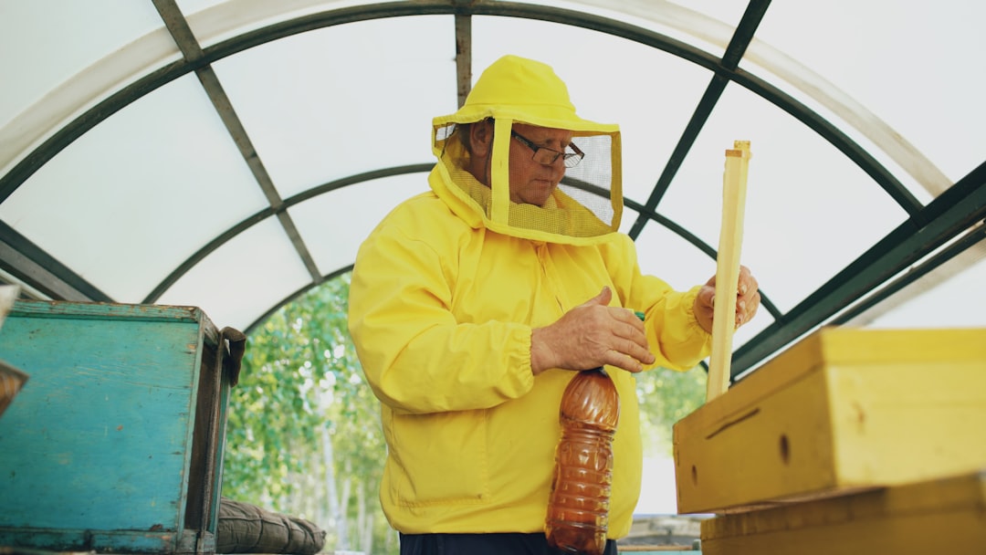 Beekeeper in full protective suit using a frame sprayer beside a wooden beehive